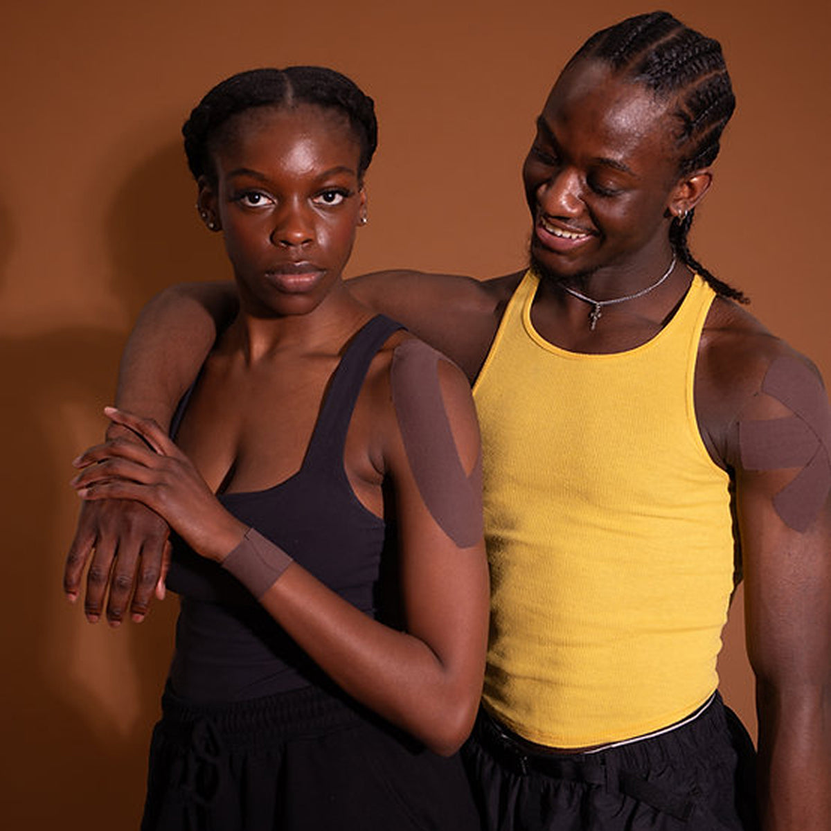 Two individuals wearing black and yellow athletic outfits with brown tape on their arms against a brown background.