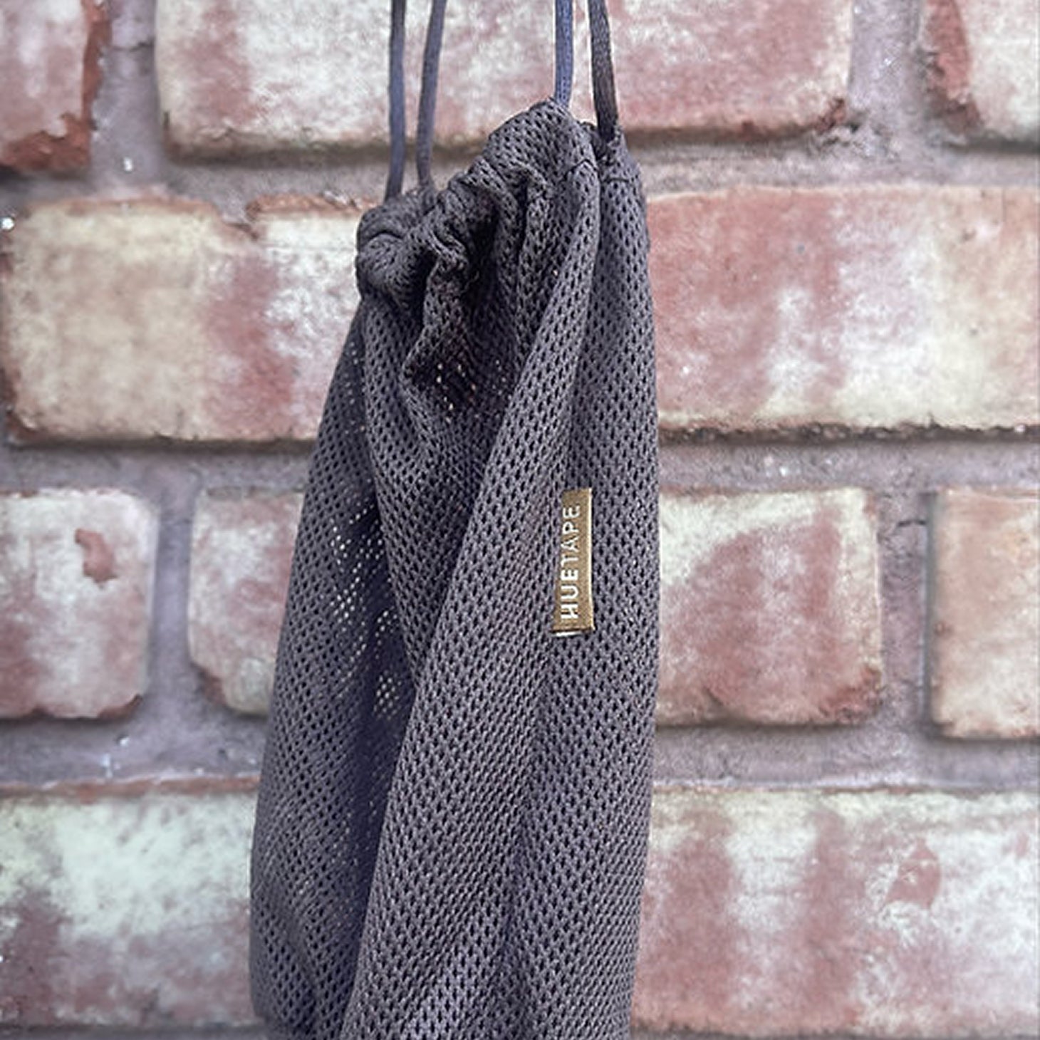 Black mesh drawstring bag with a brand label against a brick wall.